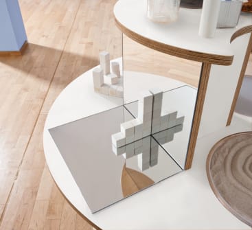 Eine weiße geometrische Skulptur aus würfelförmigen Blöcken steht auf einem runden weißen Podest neben einem quadratischen Spiegel, wobei die Skulptur und ihr kreuzförmiges Spiegelbild auf einem Holzboden sichtbar sind.