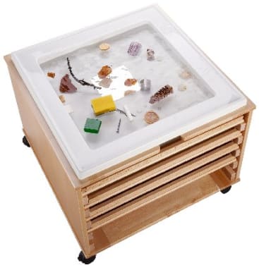 Ein hölzerner Sensoriktisch auf Rädern enthält einen durchsichtigen Behälter mit Sand, Muscheln, Blöcken, einem Kiefernzapfen und anderen kleinen Objekten, darunter befinden sich Ablageflächen.