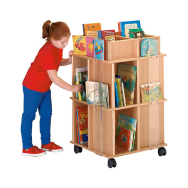 Ein junges Mädchen in rotem Hemd und blauer Hose wählt ein Buch aus einem hölzernen, mehrteiligen Bücherwagen auf Rädern aus, der mit Kinderbüchern gefüllt ist.