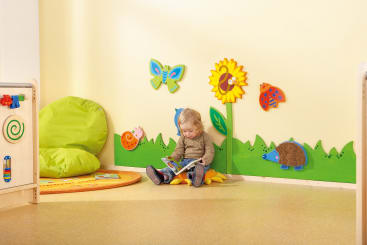 Ein kleines Kind sitzt auf einem Sitzsack und liest ein Buch in einem Spielzimmer. Die Wand dahinter ist mit bunten Tier- und Blumenausschnitten dekoriert, und ein grüner Sitzsack und Spielzeug stehen in der Nähe.