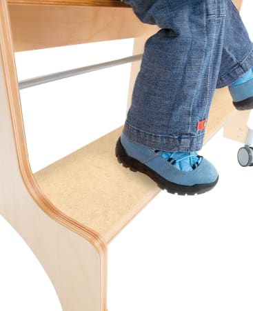 Ein Kind in blauen Turnschuhen und Jeans steht auf der Stufe eines Tritthockers aus hellem Holz und Orange, wobei ein Teil seiner Beine und Füße vor einem weißen Hintergrund zu sehen ist.