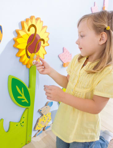 Ein junges Mädchen in einem gelben Hemd steht in einem Raum und berührt eine dekorative Sonnenblume an der Wand, die auch bunte Tier- und Insektenmotive aufweist.