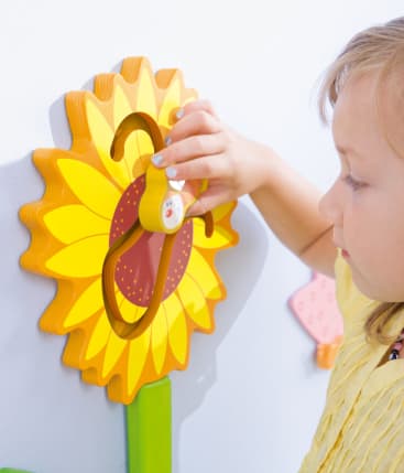 Ein kleines Kind in einem gelben Hemd bewegt eine kleine Holzbiene entlang einer Bahn auf einem an der Wand befestigten Aktivitätsbrett in Form einer Sonnenblume.