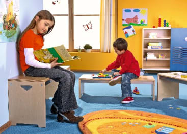 Ein Mädchen sitzt auf einer Holzbank und liest ein Buch, während ein Junge an einem niedrigen Tisch sitzt und mit Spielzeug in einem farbenfrohen Klassenzimmer mit Lernplakaten und Regalen spielt.