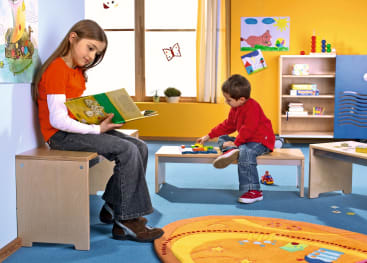 Ein Mädchen sitzt auf einer Bank und liest ein Buch, während ein Junge auf einem Tisch in einem farbenfrohen Klassenzimmer mit blauem Boden, gelben Wänden und verschiedenen pädagogischen Dekorationen und Möbeln mit Spielzeug spielt.