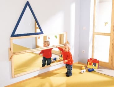 Ein Kleinkind in einem roten Hemd steht vor einem Wandspiegel und hält eine Holzstange. Spielzeug und eine Tasche liegen auf dem gelben Teppich in der Nähe. Ein großer blauer dreieckiger Rahmen hängt über dem Spiegel. Durch eine Glastür fällt natürliches Licht ein.
