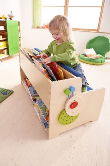 Ein kleines Kind in einem grünen Pullover blättert in Büchern in einem hölzernen Bücherregal in einem hellen, aufgeräumten Zimmer mit hellem Boden und grünem Dekor.