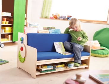 Ein kleines Kind sitzt auf einer blau gepolsterten Holzbank mit einem eingebauten Bücherregal und schaut sich ein Buch an. Auf der Bank und darunter befinden sich Kinderbücher in einem hell erleuchteten, organisierten Spielzimmer.
