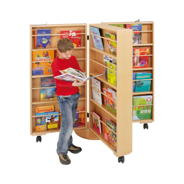 Ein Junge in rotem Hemd und Jeans steht lesend vor einem hellen, doppelseitigen, drehbaren Bücherregal auf Rädern, das mit bunten Kinderbüchern gefüllt ist.