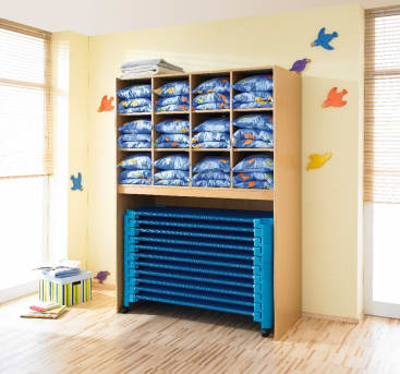 Ein hölzerner Schrank mit zwölf Fächern, gefüllt mit gefalteter blauer Bettwäsche und Handtüchern, und zehn blauen, stapelbaren Kinderbetten darunter. Der Raum hat helle Wände, die mit bunten Vogelausschnitten verziert sind.