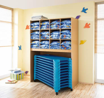 In einem hölzernen Regal steht ordentlich gefaltetes blaues Bettzeug, während darunter gestapelte blaue Kinderbetten aufbewahrt werden. An der gelben Wand in dem hellen, sonnenbeschienenen Raum mit Holzboden und großen Fenstern hängen bunte Schildkrötendekorationen.