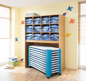 In einem Holzregal befindet sich gefaltete blaue Bettwäsche mit Cartoon-Motiven in einzelnen Fächern. Darunter sind mehrere gestapelte blaue und weiße Kinderbetten gelagert. An der hellgelben Wand sind bunte Vogeldekorationen angebracht.
