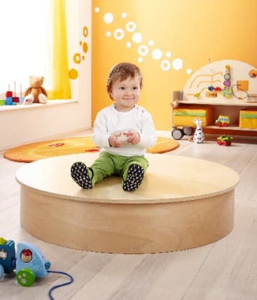Ein Kleinkind mit kurzen Haaren, einem weißen Hemd und einer grünen Hose sitzt auf einem runden Holzpodest in einem farbenfrohen Spielzimmer mit Spielzeug, einem Teppich und Wanddekorationen im Hintergrund.