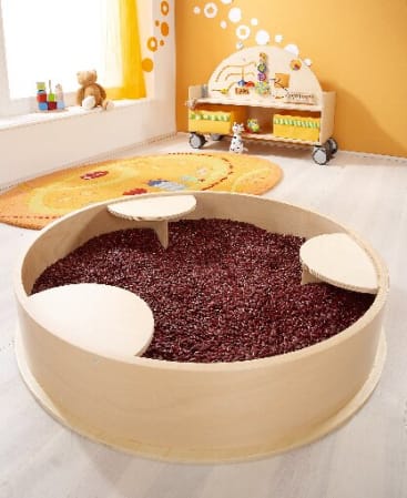 Ein hölzerner Sensoriktisch mit roten Bohnen und drei kleinen Plattformen steht in einem hell erleuchteten Spielzimmer mit einem gelben Teppich, einem niedrigen Regal mit Spielzeug und orangefarbenen, mit Kreisen verzierten Wänden.