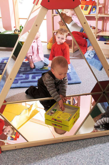 Ein kleines Kind kniet auf dem Boden und hält eine gelbe Schachtel in der Nähe einer dreieckigen Spiegelstruktur. Im Hintergrund eines Spielzimmers sind zwei andere Kinder und Spielzeug zu sehen.