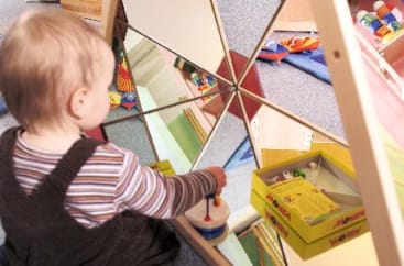 Ein kleines Kind in gestreifter Kleidung sitzt vor einer Spiegelwandstruktur und hält ein Spielzeug in der Hand. In der Spiegelung und im Hintergrund sind eine gelbe Schachtel mit Mosaikstiften und anderes Spielzeug zu sehen.