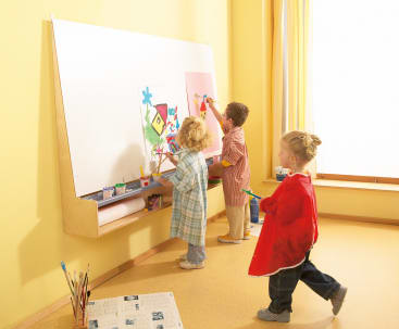 Drei kleine Kinder malen auf einer großen weißen Tafel, die an einer gelben Wand befestigt ist. Auf einem Sims unterhalb der Tafel stehen Malutensilien und Farbdosen. Das Sonnenlicht fällt durch ein Fenster mit gelben Vorhängen auf der rechten Seite.