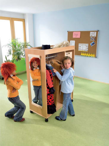 Zwei Kinder mit orangefarbenen Perücken und Jacken stehen an einem hölzernen Kostümschrank mit einem Spiegel, während ein drittes Kind mit einer blauen Jacke sie ansieht. Der Raum hat einen grünen Fußboden und eine Pinnwand an der Wand.