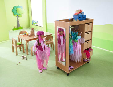 Ein kleines Kind in einem rosafarbenen Feenkostüm betrachtet sein Spiegelbild in einem hölzernen Schrank, der mit bunten Kostümen gefüllt ist. Der Raum hat grüne Wände, Tische und Stühle in Kindergröße und ein Wandbild mit einem Baum.