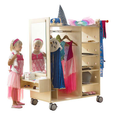 Ein kleines Kind steht vor einem hölzernen Verkleidungswagen mit einem Spiegel und trägt ein rosa Kleid und ein Stirnband. Auf dem Wagen befinden sich Kostüme, Hüte, ein Zaubererhut und anderes Zubehör, mit Regalen und Aufhängemöglichkeiten.