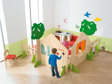 Zwei kleine Kinder spielen in einem hell erleuchteten Spielzimmer mit hölzernen Spielgeräten in Form von Häusern und Bäumen. Der Raum hat grüne und weiße Wände, einen Holzboden und Spielzeugküchenmöbel im Hintergrund.