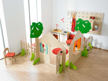 Ein Spielbereich für Kinder mit hölzernen Spielgeräten in Form eines Hauses und von Bäumen, einem kleinen Küchenset, einem Stuhl mit rotem Kissen, einem Teddybär und einem hellen Holzboden. Rote und orangefarbene Akzente sind überall zu sehen.
