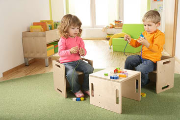 Zwei kleine Kinder sitzen auf Holzstühlen an einem kleinen Tisch und fädeln bunte Perlen auf Schnüre auf. Im Hintergrund sind Regale mit Büchern und Spielzeug sowie eine grüne Couch auf einem grünen Teppichboden zu sehen.