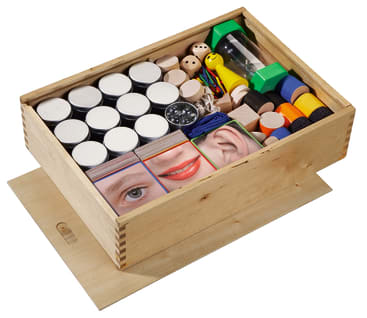 Eine Holzkiste mit Schiebedeckel enthält Teelichter, Holzteile, Karten mit Teilen eines Gesichts, eine Sanduhr, einen Kompass, gelbe und grüne Figuren und verschiedene strukturierte Gegenstände.