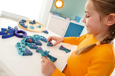 Ein Kind in einem orangefarbenen Hemd ordnet bunte, mit Edelsteinen verzierte Holzpuzzleteile auf einem weißen Tisch an. Weitere Teile und ein Holztablett sind in der Nähe zu sehen. Im Hintergrund sind ein Schrank und eine grüne Wand zu sehen.