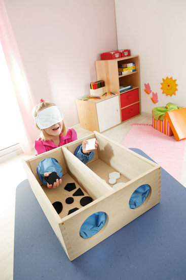 Ein kleines Kind mit einer Augenbinde sitzt an einem Tisch und greift in eine hölzerne Sinnesbox mit verschieden geformten Löchern und taktilen Objekten. Der Raum ist hell mit Kindermöbeln und bunten Dekorationen.