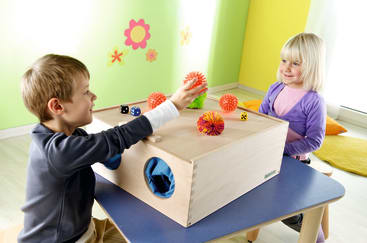 Zwei kleine Kinder sitzen an einem blauen Tisch und spielen mit stacheligen Sinneskugeln und Würfeln auf einer hölzernen Sinnesbox. Der Raum hat leuchtend gelbe Wände mit Blumenaufklebern und bunte Kissen auf dem Boden.