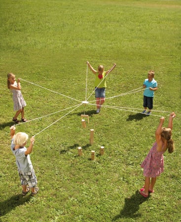 Fünf Kinder stehen im Kreis auf dem Rasen, jedes hält eine Schnur, die mit einem zentralen Ring verbunden ist, und hebt oder stellt Holzklötze in der Mitte der Gruppe aufrecht.