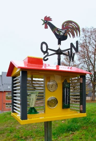 Eine gelb-rote Wetterstation mit einer Wetterfahne in Form eines Hahns auf der Spitze, mit Messgeräten und einem Thermometer, steht draußen auf einer Wiese in der Nähe von Gebäuden und Bäumen.