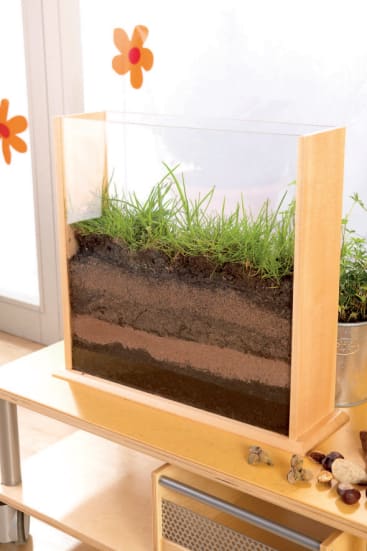 Eine rechteckige Glasvitrine zeigt Schichten von Erde, Sand und Ton, auf denen Gras wächst. Die Vitrine steht auf einem hellen Holztisch neben einem Metalltopf und einigen kleinen Steinen. An einem Fenster in der Nähe sind orangefarbene Blumenaufkleber angebracht.