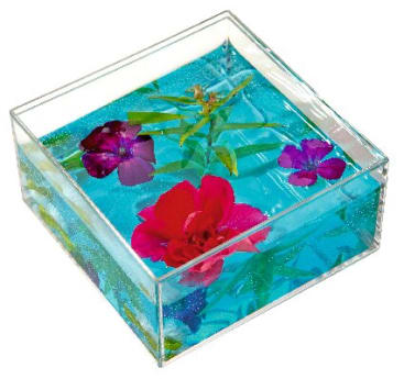 Ein durchsichtiger, quadratischer Behälter, gefüllt mit klarem, blauem Gel, in dem mehrere bunte Kunstblumen und grüne Blätter angeordnet sind.