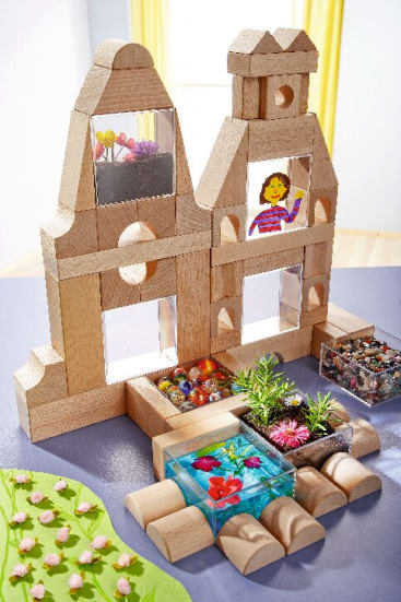 Eine Struktur aus Holzbausteinen mit transparenten Teilen enthält kleine Dekorationen. Davor stehen Tabletts mit bunten Perlen, Pflanzen und Spielzeugfischen. In einem Fenster befindet sich eine gezeichnete Figur. Im Hintergrund ist ein gelber Vorhang zu sehen.