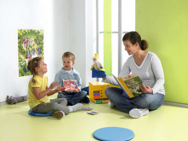 Eine Frau sitzt mit zwei kleinen Kindern in einem hell erleuchteten Raum auf dem Boden und hält Bücher in der Hand. Die Kinder sitzen auf runden Matten und schauen sie an, und auf dem Boden liegen ein Spielzeug und Bildkarten. Ein buntes Poster hängt an der Wand.