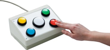 Die Hand einer Person drückt einen roten Knopf auf einem weißen, rechteckigen Schaltkasten mit fünf großen farbigen Tasten: gelb, grün, weiß, blau und rot. Der Kasten ist mit einem Kabel verbunden.