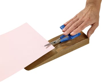 Eine Hand hält eine blaue Schere auf einem Holzsockel, während sie ein Blatt rosa Papier ausschneidet. Die Anordnung hilft beim gleichmäßigen, einhändigen Schneiden.