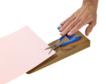 Eine Hand benutzt eine blaue adaptive Schere auf einem Holzsockel, um ein Blatt hellrosa Papier zu schneiden.