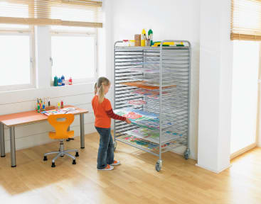 Ein Kind steht in einem hellen Klassenzimmer und legt ein Bild auf ein Trockengestell, das mit Kunstwerken gefüllt ist. Auf der linken Seite stehen ein Schreibtisch mit Malutensilien und ein orangefarbener Stuhl. Der Raum hat Holzböden und große Fenster mit Jalousien.