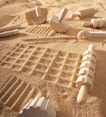 Auf einer sandigen Fläche sind hölzerne Sandrollen und Strukturwerkzeuge verstreut, die verschiedene Muster und Linien in den Sand eingedrückt haben. Die Werkzeuge sind zylindrisch und haben verschiedene Rillenmuster.