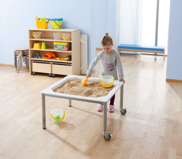 Ein Kleinkind spielt mit einer Schaufel mit Sand an einem sensorischen Tisch in einem Klassenzimmer mit Holzregalen, Hockern und verschiedenen Spielmaterialien im Hintergrund.