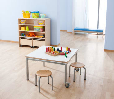Ein Klassenzimmer mit hellem Holzboden, einem kleinen Tisch mit zwei runden Holzhockern, bunten Holzklötzen auf dem Tisch und einem Regal mit Behältern und Spielzeug im Hintergrund. Licht strömt durch ein Fenster herein.