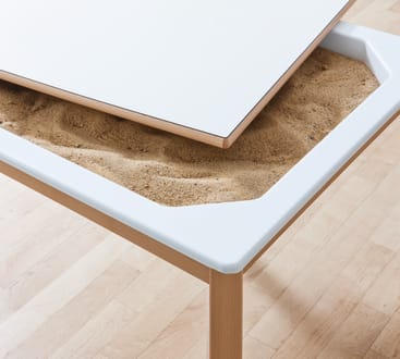 Nahaufnahme eines Tisches mit einer teilweise angehobenen weißen Tischplatte, die eine Sandschicht in einem vertieften Fach unter der Oberfläche erkennen lässt. Der Tisch hat helle Holzbeine und steht auf einem Holzboden.