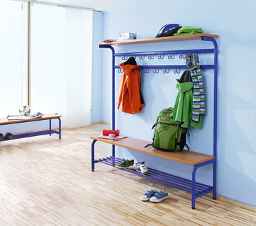 Eine Garderobe mit einer Holzbank und einem Regal, auf dem Jacken, ein grüner Rucksack, Mützen und Schals liegen. Auf dem Boden und der Bank stehen Schuhe, eine Wasserflasche und ein Federmäppchen. Der Raum hat einen hellen Holzfußboden und große Fenster.