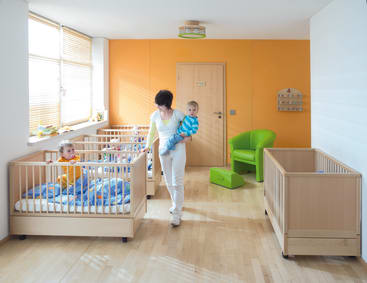 Eine Frau trägt ein Kind, während sie an Krippen in einem hell erleuchteten Kinderzimmer mit orangefarbenen und weißen Wänden, Holzböden und einem grünen Stuhl vorbeigeht. Ein anderes Kind sitzt in einer Krippe.