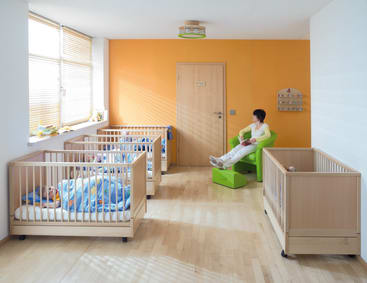 Ein Kinderbetreuungsraum mit drei Holzbetten, in denen jeweils ein Kind schläft. Eine Frau sitzt auf einem grünen Stuhl in der Nähe der Tür und schaut den Kindern zu. Die Wände sind orange und weiß, und der Boden ist aus hellem Holz.