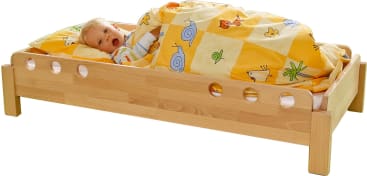 Ein kleines Kind liegt in einem Holzbett, das mit einer gelben und orangefarbenen, mit Tieren und Schnecken verzierten Decke bedeckt ist, und blickt mit offenem Mund nach oben. Das Bett hat kreisförmige Ausschnitte in den Seitenwänden.