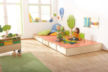 Zwei kleine Kinder spielen mit buntem Bauspielzeug auf einem niedrigen Podest in einem hellen, aufgeräumten Raum mit Holzböden, großen Fenstern und verschiedenen Dekorationen und Lernspielzeug an den Wänden und in den Regalen.